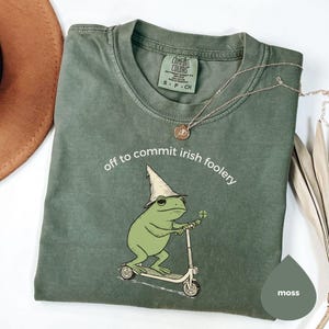 May include: Moss green t-shirt with the phrase "off to commit irish foolery" and a graphic of a frog riding a scooter, wearing a wizard hat and holding a four-leaf clover. A gold necklace is draped across the shirt.