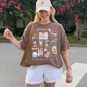 Może przedstawiać: Brązowa koszulka z grafiką Disney World z Myszką Miki, Minnie, Kaczorem Donaldem i innymi postaciami. Koszulka ma napis "Walt Disney World" i "2026". Osoba nosi białe szorty i beżową czapkę z daszkiem.