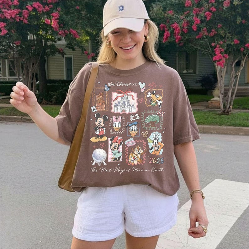 Może przedstawiać: Brązowa koszulka z grafiką Disney World z Myszką Miki, Minnie, Kaczorem Donaldem i innymi postaciami. Koszulka ma napis "Walt Disney World" i "2026". Osoba nosi białe szorty i beżową czapkę z daszkiem.