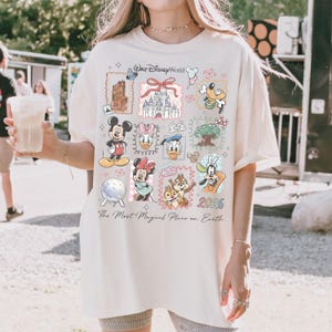 Może przedstawiać: Kremowa koszulka z kolażem postaci i zabytk&oacute;w Disneya. Projekt zawiera Myszkę Miki, Minnie, Kaczora Donalda i Goofy'ego, wraz z tekstem "Walt Disney World" i "2026". Koszulka zawiera r&oacute;wnież frazę "The Most Magical Place on Earth".