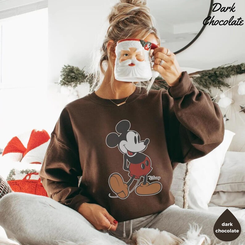 May include: A dark chocolate-coloured sweatshirt featuring a smiling Mickey Mouse graphic. The sweatshirt has a crew neck and long sleeves. The image also shows a Santa Claus mug and the text "Dark Chocolate".