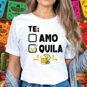 May include: White t-shirt with the text "TE: AMO QUILA" in black. The design includes a checkmark next to "QUILA" and an illustration of a tequila shot with a lime wedge and mint leaves. Festive decorations are in the background.