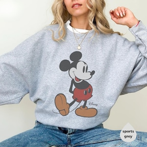 May include: A light grey sweatshirt with a Mickey Mouse graphic. The cartoon character is shown in his classic pose, wearing red shorts and brown shoes. The Disney logo is printed below the character. The sweatshirt is sports grey.
