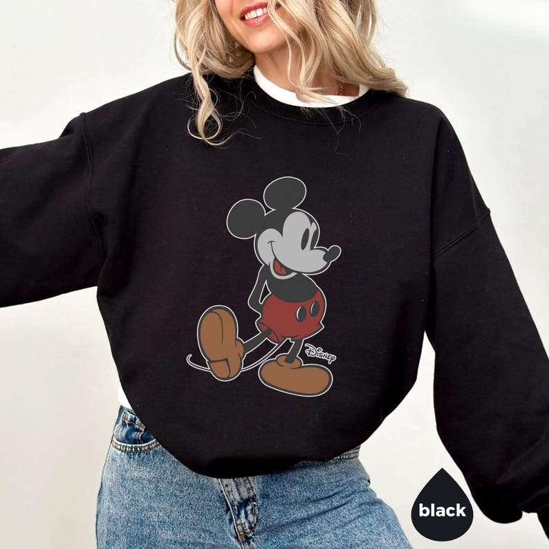 May include: Black sweatshirt with a Mickey Mouse graphic. The cartoon character is shown in his classic pose, wearing red shorts and brown shoes. The word "Disney" is printed on the bottom right of the graphic. The sweatshirt has a crew neck and long sleeves.