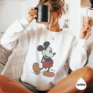 May include: White sweatshirt with a Mickey Mouse graphic. The cartoon character is shown in his classic pose, wearing red shorts and brown shoes. The Disney logo is present. The sweatshirt is a casual style.