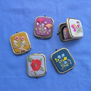 May include: Five square compact mirrors with embroidered fabric covers. The designs feature floral motifs in yellow, purple, white, light gray, and blue fabric. Each mirror has a metallic frame and displays colorful stitched flower patterns.