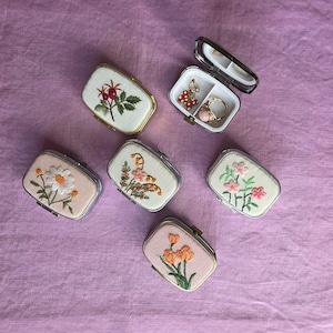 May include: A collection of small, rectangular embroidered jewelry boxes with floral designs. One box is open, revealing jewelry. The boxes feature various flower patterns in shades of pink, orange, and white, set against a neutral background.