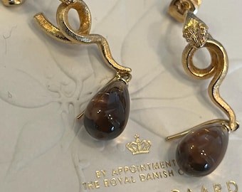 May include: A pair of gold-toned snake earrings with brown teardrop-shaped pendants. The earrings feature detailed snake heads and bodies, with a textured finish. The background includes text that reads "OLE LYNGGAARD" and "SINCE 1963".