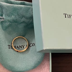 Può includere: Un anello d'oro con piccoli accenti rossi poggia su un sacchetto turchese con le parole "TIFFANY & Co." stampate sopra. Una scatola azzurra con la parola "TIF" visibile è aperta accanto al sacchetto.