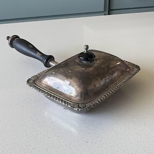 Op de afbeelding: Een rechthoekige verzilverde serveerschaal met deksel, voorzien van een sierlijke decoratieve rand, een donkerhouten handvat en een kleine zwarte knop op het deksel. Het metalen oppervlak vertoont tekenen van aanslag en slijtage.