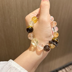 以下が含まれることがあります： 黄色、茶色、透明、濃い灰色の様々な丸い天然石ビーズで構成された伸縮性のあるブレスレット。ビーズは、滑らかに磨かれた表面、ファセットカット、石内部のインクルージョンなど、多様な質感と特徴を備えています。