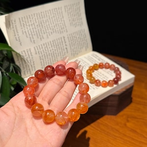 May include: Two beaded bracelets made of translucent, spherical stones in shades of orange, red, and deep crimson. The beads feature internal inclusions and a polished finish. One bracelet is held in the foreground, while the other rests on an open book.