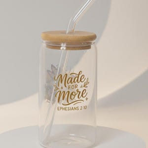 Puede incluir: Un vaso de vidrio transparente con tapa de bambú y pajita transparente. El vaso tiene la inscripción dorada "Made for More" y "Ephesians 2:10". También se aprecia un diseño floral.