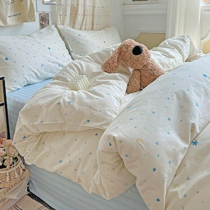 Puede incluir: Juego de cama color crema con un patrón celestial de estrellas azules y lunas crecientes. El juego incluye una funda nórdica, fundas de almohada y una sábana bajera. Un juguete de peluche de perro marrón descansa sobre la cama.