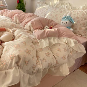 Puede incluir: Una cama con un edredón y fundas de almohada con estampado floral en tonos rosa y crema. El edredón tiene una capa superior rosa y ribete de volantes blancos. Un conejito de peluche con un sombrero azul está en la cama.