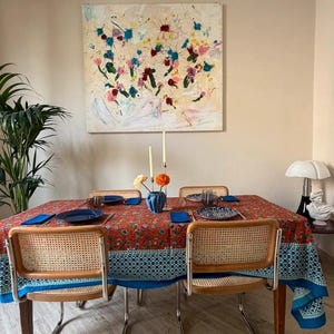 May include: A dining room scene featuring a table set with a red floral tablecloth and four chairs. The chairs have woven backs and chrome legs. A painting with colorful splatters hangs on the wall above the table.