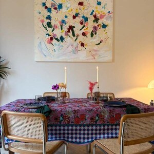 Può includere: Una scena di sala da pranzo con un tavolo apparecchiato. Il tavolo è coperto da una tovaglia a fantasia floreale. Sono visibili due sedie in rattan. Un dipinto con fiori astratti colorati è appeso alla parete sopra il tavolo.
