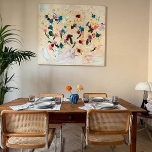 Può includere: Una sala da pranzo con un tavolo di legno apparecchiato per quattro persone, con sedie con schienale in vimini. Un dipinto astratto con spruzzi colorati è appeso sopra. Un vaso blu con fiori è sul tavolo.