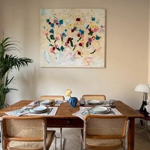 Può includere: Una sala da pranzo con un tavolo di legno apparecchiato per quattro persone. Il tavolo è adornato con tovagliette, piatti e bicchieri. Sedie in vimini con telai in metallo circondano il tavolo. Un dipinto astratto con pennellate colorate è appeso al muro.