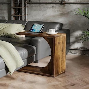 May include: A wooden side table with a dark brown finish, featuring a unique curved design on one side. The table is positioned next to a gray leather sofa, with a tablet and a cup of coffee on top. The table is approximately 24 inches tall.