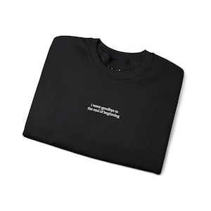May include: Black crew neck sweatshirt with the text "i wave goodbye to the end of beginning" in white. The sweatshirt is folded neatly on a white surface. The text is centered on the chest.
