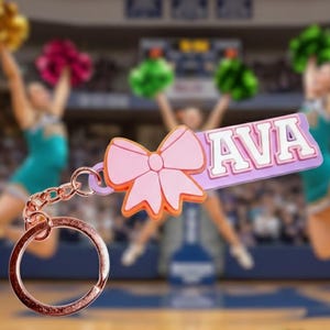 May include: A pink bow-shaped keyring with the name "AVA" in white letters on a purple background. The keyring has a rose gold ring and chain. Cheerleaders with pom-poms are in the background.
