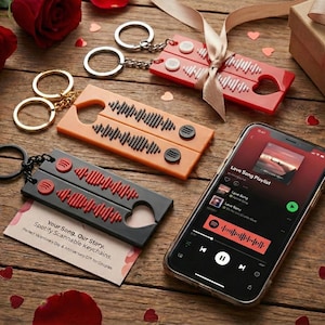 Pode incluir: Chaveiros com design inspirado no Spotify em vermelho, laranja e preto, juntamente com um smartphone exibindo uma lista de reprodução. Os chaveiros têm um padrão de onda sonora e recortes em forma de coração. Rosas e uma caixa de presente também estão presentes.