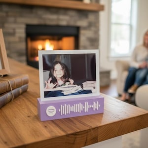 May include: A framed photo of a smiling girl sits on a lavender Spotify code stand. A framed dog portrait and two antique books are also on the wooden table. The background shows a fireplace and a family.