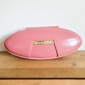 May include: A pink plastic sewing machine case with the word "SINGER" in gold lettering on the front.