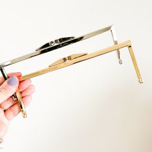 May include: Two metal purse frames, one silver and one gold, are held in a hand. The frames are rectangular with a rounded top and bottom.