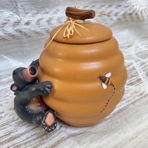 Könnte beinhalten: Ein Keramik-Honigtopf in Form eines Bienenstocks mit beiger Oberfläche und einer seitlich angebrachten braunen Bärenfigur. Der Deckel hat einen braunen Griff und ein Bastband. Eine kleine gemalte Biene mit Flugbahn ist vorne zu sehen.