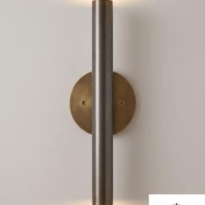 May include: A wall-mounted light fixture featuring a long, cylindrical, dark metallic tube centered over a circular, brushed brass backplate. The fixture emits a soft, warm glow from both the top and bottom openings of the central tube.