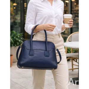 May include: A navy blue leather handbag with a structured design, featuring top handles and a detachable shoulder strap. The bag has gold-toned hardware and a rectangular shape. The person is wearing a white button-down shirt and tan pants.