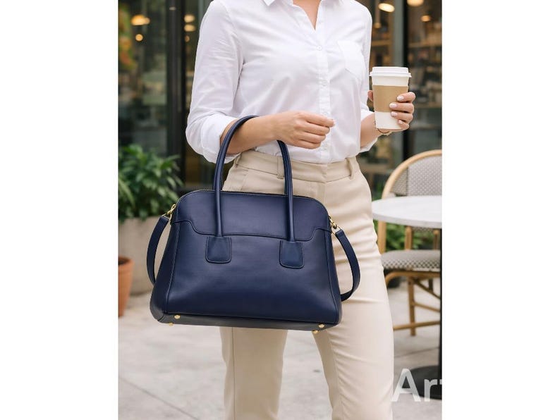 May include: A navy blue leather handbag with a structured design, featuring top handles and a detachable shoulder strap. The bag has gold-toned hardware and a rectangular shape. The person is wearing a white button-down shirt and tan pants.