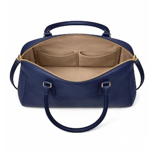 May include: Open navy blue leather handbag with gold-tone hardware. The interior features two pockets and a beige lining. The bag has two handles and a zipper closure.