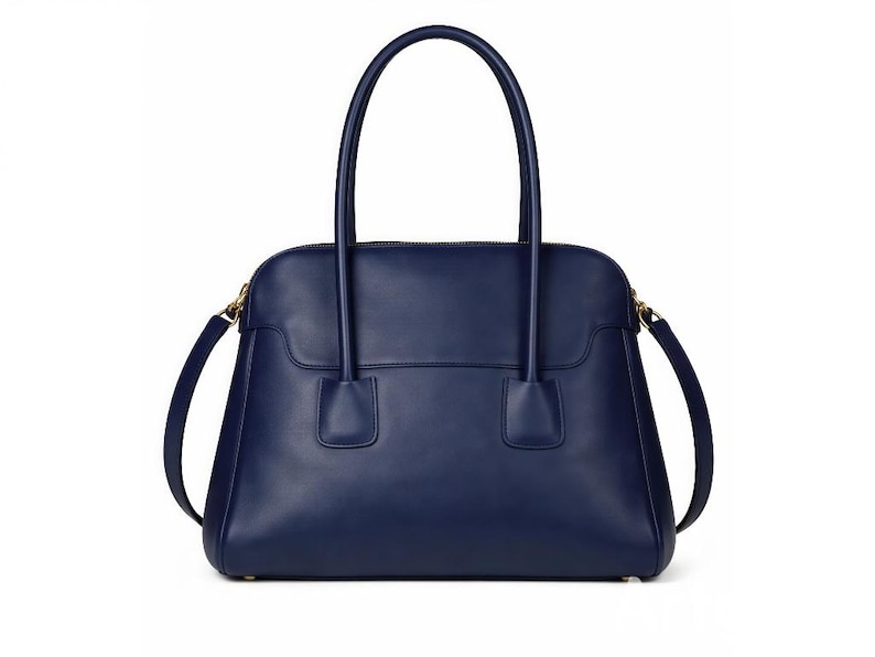 May include: A navy blue leather handbag with a structured design. The bag features two top handles and a detachable shoulder strap. Gold-toned hardware accents the zipper and strap attachments. The bag is approximately 12 inches wide.