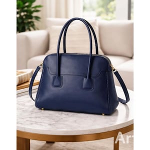 May include: A navy blue leather handbag with a structured design. The bag features a top handle, a detachable shoulder strap, and gold-toned hardware. The bag is sitting on a marble table.