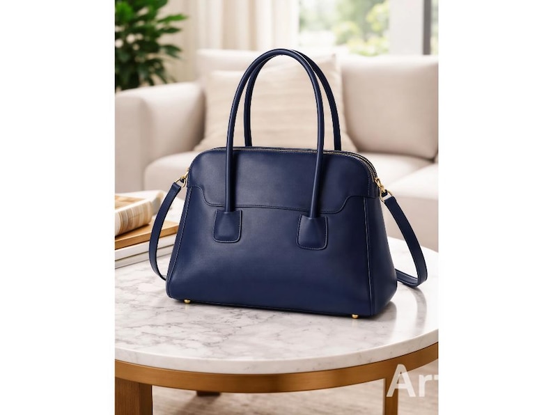 May include: A navy blue leather handbag with a structured design. The bag features a top handle, a detachable shoulder strap, and gold-toned hardware. The bag is sitting on a marble table.