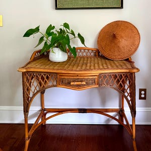 May include: A framed artwork depicting a seated figure in gold tones, hanging above a bamboo desk. The desk features a drawer and a woven top, with a white vase holding green plants. A woven hat rests on the desk.