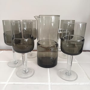 May include: A set of vintage glassware featuring a pitcher and several wine glasses. The glasses and pitcher have a smoky gray color on the bottom half, with clear stems and bases. The glassware is arranged on a white tiled surface.