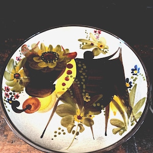 May include: A white ceramic plate with a hand-painted design of a black bull with yellow and green floral accents.