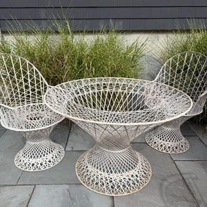 May include: Outdoor patio set with a round table and two matching chairs. The furniture is made of white woven material, featuring a unique open design. The table and chairs are placed on a stone patio with greenery in the background.