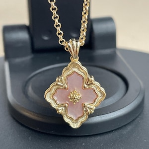 Op de afbeelding: Een goudkleurige ketting met een hanger in de vorm van een klavertjevier, met een roze centrum en een goudkleurige rand. De hanger heeft een kleine, goudkleurige bloem in het midden. De ketting is ook goudkleurig.