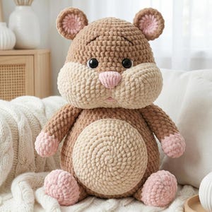 May include: A handmade, plush crochet hamster toy in shades of brown, tan, and pink. The stuffed animal has large cheeks, small ears, and a round belly. The hamster is sitting on a white blanket.