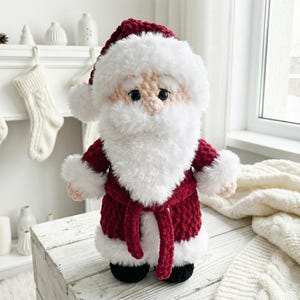 May include: A handmade crochet Santa Claus doll. The doll features a red suit with white trim, a matching hat, and a fluffy white beard. The doll is standing on a white wooden surface. The doll is a festive decoration.