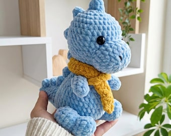 May include: A light blue crocheted dinosaur toy with a yellow scarf. The plush dinosaur has black eyes and is made of a soft, textured yarn. The toy is approximately 6 inches tall and is a handmade item.