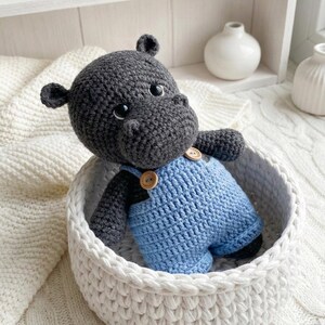 May include: A handmade crochet hippo toy in a white basket. The hippo is gray with black accents and wears blue overalls with wooden buttons. The basket is made of thick, white yarn.