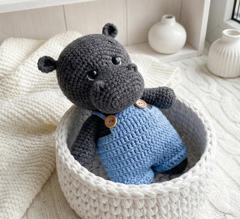 May include: A handmade crochet hippo toy in a white basket. The hippo is gray with black accents and wears blue overalls with wooden buttons. The basket is made of thick, white yarn.