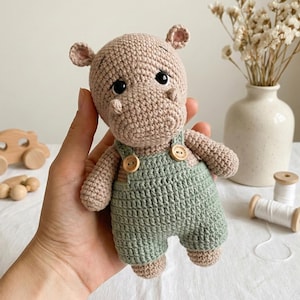 ALT TEXT (5)
1) Crochet hippo doll with beige yarn body and black safety eyes, wearing green overalls with two wooden-style buttons, held in hands.
2) Close-up of amigurumi hippo face showing rounded snout shaping, tiny ears, and glossy safety eyes.
3) Front view of crochet hippo plush in green overalls, showing strap details and button accents on the chest.
4) Nursery-style photo of the crocheted hippo toy held in hands with cozy neutral decor, wooden toys, and dried florals in the background.