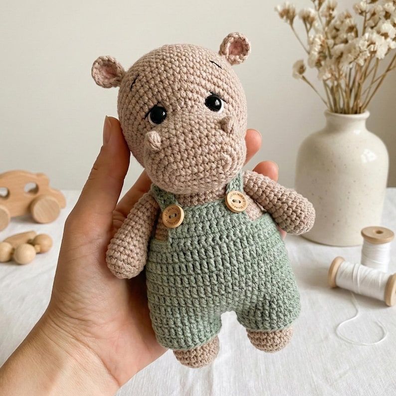 ALT TEXT (5)
1) Crochet hippo doll with beige yarn body and black safety eyes, wearing green overalls with two wooden-style buttons, held in hands.
2) Close-up of amigurumi hippo face showing rounded snout shaping, tiny ears, and glossy safety eyes.
3) Front view of crochet hippo plush in green overalls, showing strap details and button accents on the chest.
4) Nursery-style photo of the crocheted hippo toy held in hands with cozy neutral decor, wooden toys, and dried florals in the background.